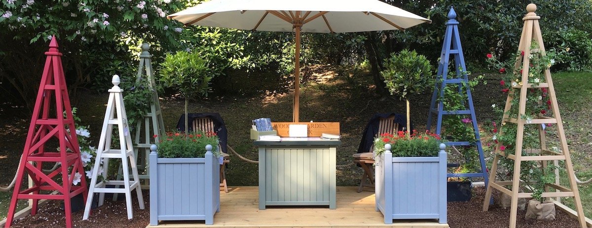 Wooden Garden Obelisk stand at RHS Chelsea Flower Show 2018 includes a pair of Versailles Style Planters painted Manor House Gray, each planted with specimen Bay Trees. The left side includes Chelsea Star Obelisk painted Rectory Red by Farrow & Ball, 160cm Wooden Garden Obelisk painted Lime White by Farrow & Ball, 195cm Sweet Pea Obelisk painted Lichen by Farrow & Ball. Right side  of stand includes a 230cm Wooden Garden Obelisk painted Juniper Ash by Little Greene and a 230cm Oak Garden Obelisk